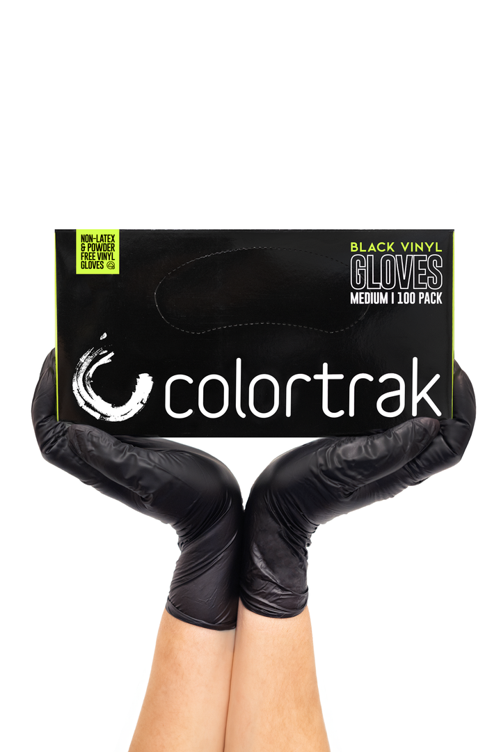 Two gloved hands holding a black box of Colortrak black vinyl gloves with a lime green label, 100 pack