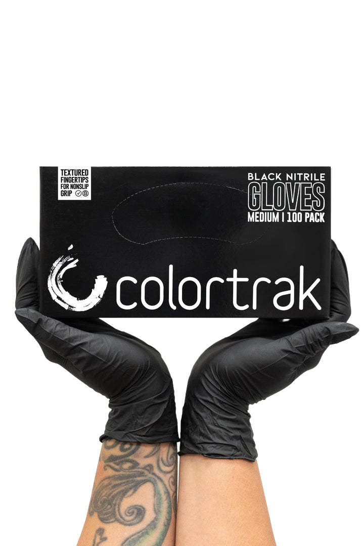 Two gloved hands holding a black box of Colortrak Midnight Black nitrile gloves, 100 pack
