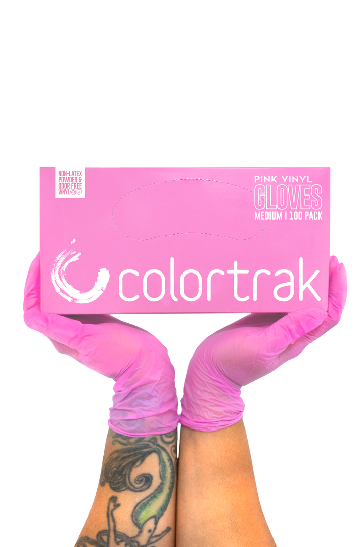 Two gloved hands holding a pink box of Colortrak pink vinyl gloves, 100 pack