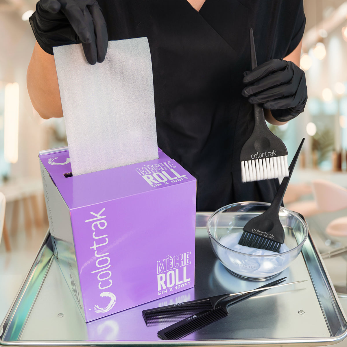 Stylist in black gloves pulls meche from the Colortrak Mèche Roll box, 5 in x 100 ft, on a silver tray with two color brushes, a clear bowl of lavender lightener and two tail combs.