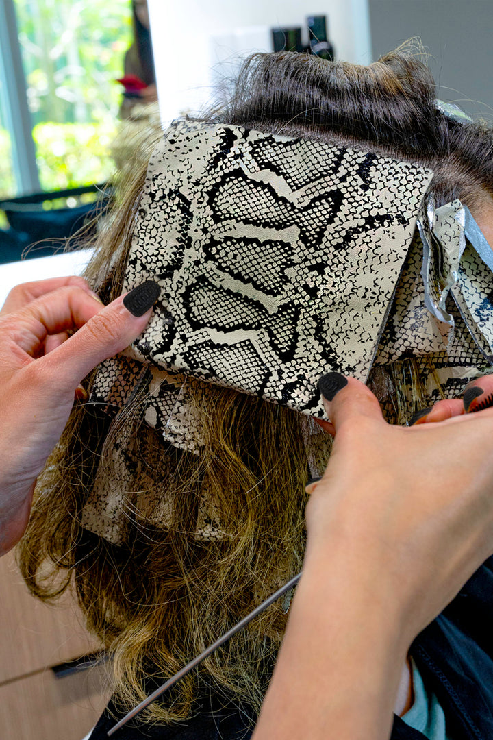 Hands folding a Python print foil into a section of brown hair during a color service